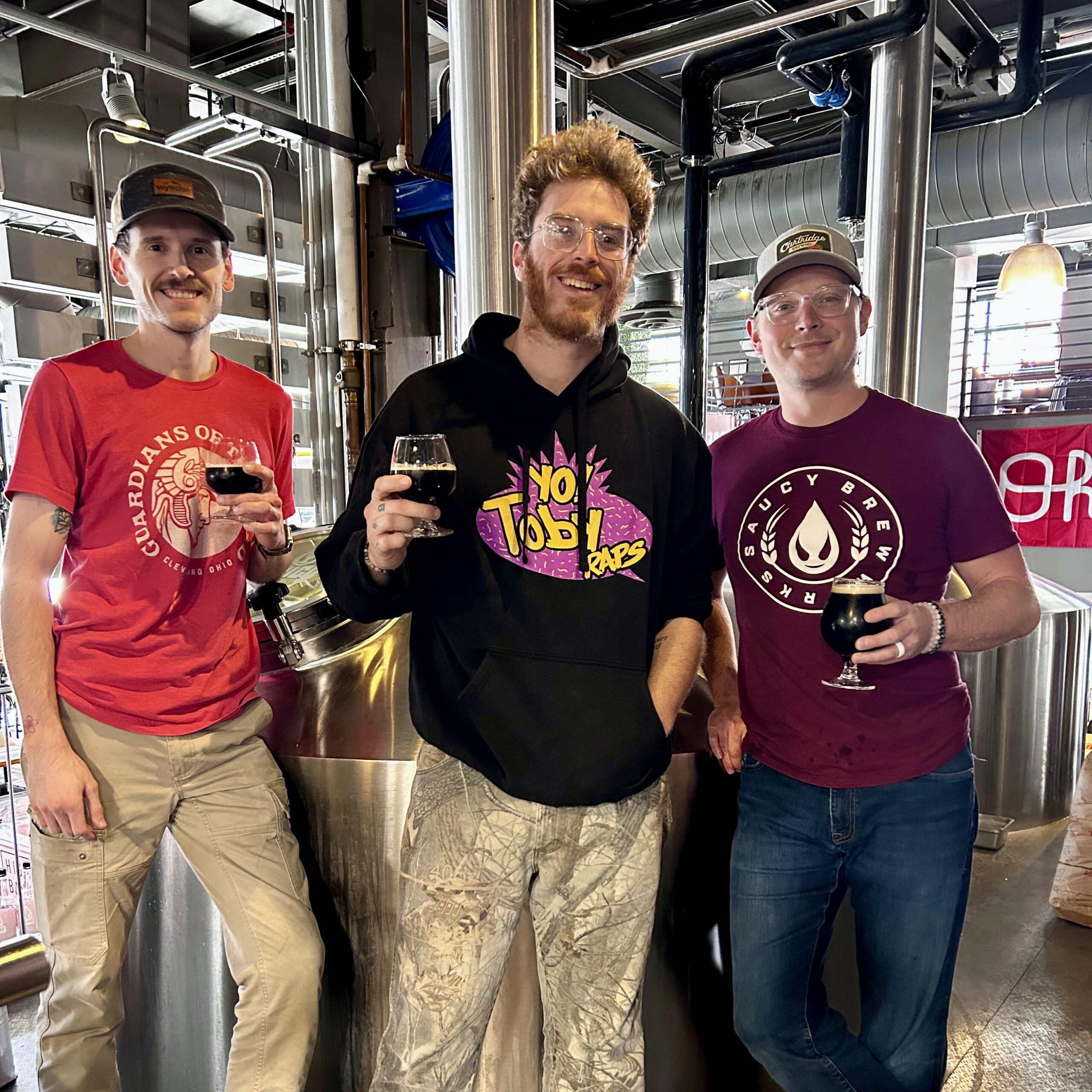 Tobyraps and two brewers holding beers surrounded by brewing tanks.