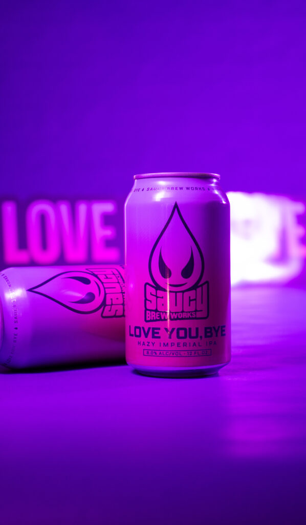 Two cans of Loe You, Bye in front of a Love You, Bye sign on a glowing purple background.